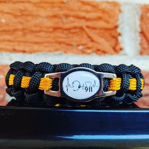 May include: Black and yellow paracord bracelet with a silver charm featuring a stethoscope, heart, and the number 911.
