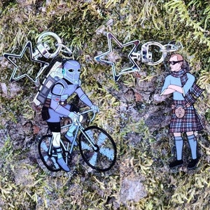 May include: Two keychains featuring a silver star design. One keychain has a blue and gray figure riding a bicycle, while the other keychain has a blue figure wearing a kilt.