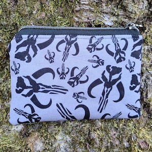 Puede incluir: Un peque&ntilde;o monedero rectangular con cierre de cremallera negro. La tela gris claro est&aacute; estampada con s&iacute;mbolos de calaveras mandalorianas negras. El monedero est&aacute; sobre un fondo de madera musgosa y texturizada.