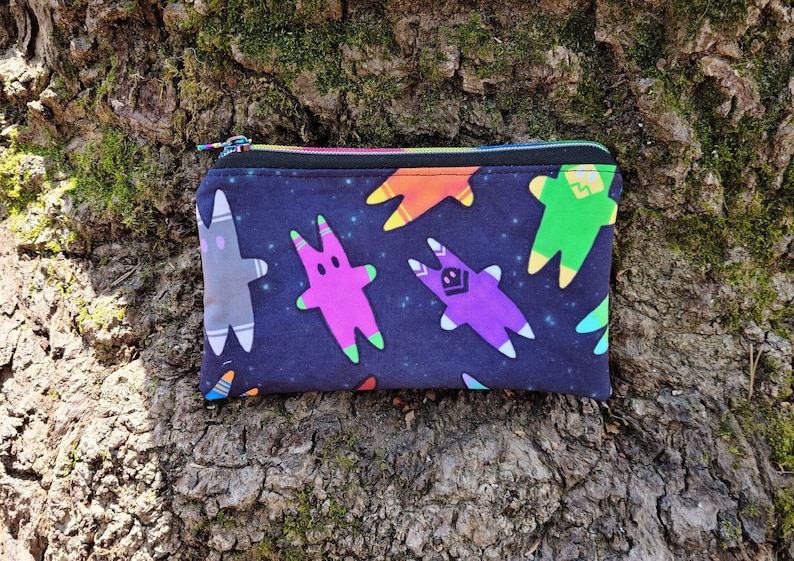 Puede incluir: Una bolsa negra con cremallera con un estampado colorido de tem&aacute;tica espacial con varios personajes de dibujos animados. Los personajes son de diferentes colores y tienen forma de estrella.