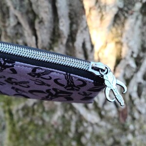 Puede incluir: Primer plano de un estuche morado con un patr&oacute;n negro y una cremallera plateada. El tirador de la cremallera es un dije plateado. El fondo es un tronco de &aacute;rbol borroso.