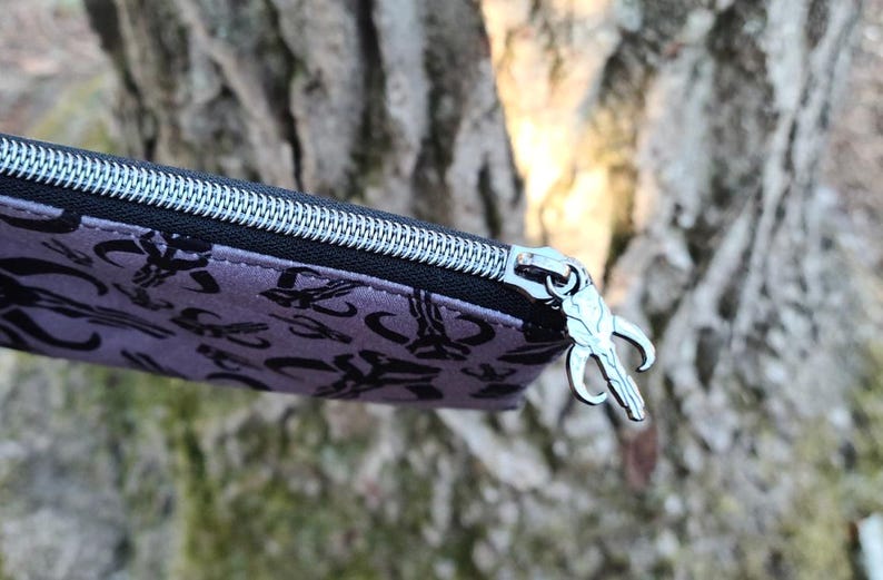 Puede incluir: Primer plano de un estuche morado con un patr&oacute;n negro y una cremallera plateada. El tirador de la cremallera es un dije plateado. El fondo es un tronco de &aacute;rbol borroso.