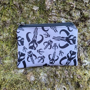 Puede incluir: Una peque&ntilde;a bolsa rectangular con cremallera, con un fondo gris claro y un patr&oacute;n gr&aacute;fico negro repetido. La bolsa est&aacute; sobre una superficie musgosa y texturizada. La cremallera es negra y plateada.