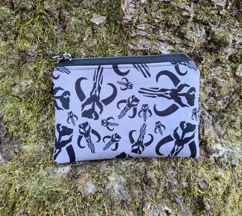 Puede incluir: Una peque&ntilde;a bolsa rectangular con cremallera, con un fondo gris claro y un patr&oacute;n gr&aacute;fico negro repetido. La bolsa est&aacute; sobre una superficie musgosa y texturizada. La cremallera es negra y plateada.