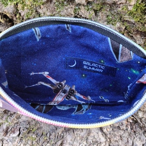 Puede incluir: Una bolsa negra con cremallera con un tirador de cremallera arco&iacute;ris. El interior de la bolsa est&aacute; forrado con una tela azul con un estampado de tem&aacute;tica espacial con estrellas, planetas y naves espaciales. La tela tambi&eacute;n tiene una etiqueta negra con el texto "GALACTIC SUMMER".