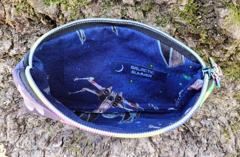 Puede incluir: Una bolsa negra con cremallera con un tirador de cremallera arco&iacute;ris. El interior de la bolsa est&aacute; forrado con una tela azul con un estampado de tem&aacute;tica espacial con estrellas, planetas y naves espaciales. La tela tambi&eacute;n tiene una etiqueta negra con el texto "GALACTIC SUMMER".