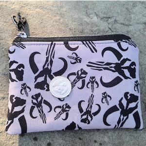 Puede incluir: Un peque&ntilde;o monedero rectangular con cremallera negra y cierre plateado. La tela morada clara est&aacute; estampada con s&iacute;mbolos de calaveras mandalorianas negras. Una moneda plateada descansa sobre el monedero.