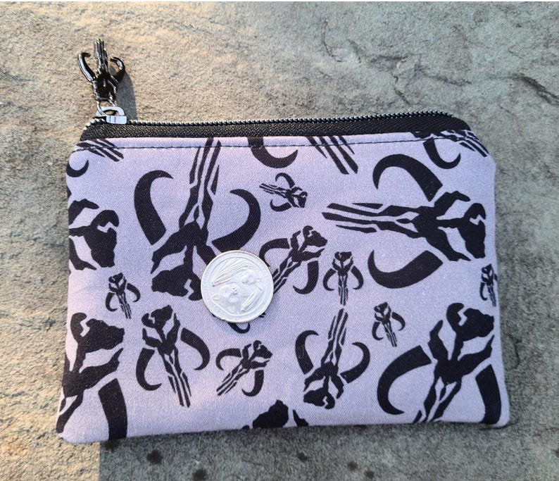 Puede incluir: Un peque&ntilde;o monedero rectangular con cremallera negra y cierre plateado. La tela morada clara est&aacute; estampada con s&iacute;mbolos de calaveras mandalorianas negras. Una moneda plateada descansa sobre el monedero.