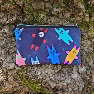 Puede incluir: Una bolsa negra con cremallera con un estampado colorido de personajes de dibujos animados sobre un fondo azul oscuro. Los personajes son de varios colores, incluyendo rosa, azul, amarillo y verde.