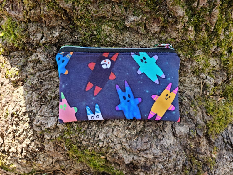 Puede incluir: Una bolsa negra con cremallera con un estampado colorido de personajes de dibujos animados sobre un fondo azul oscuro. Los personajes son de varios colores, incluyendo rosa, azul, amarillo y verde.