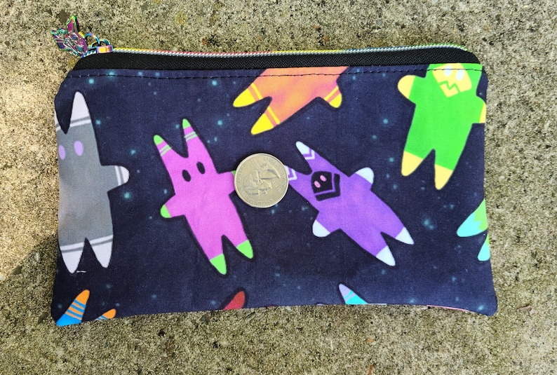 Puede incluir: Una bolsa negra con cremallera con un estampado de tem&aacute;tica espacial con figuras de dibujos animados coloridas sobre un fondo azul oscuro con estrellas blancas.