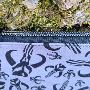 Puede incluir: Peque&ntilde;o estuche rectangular con cremallera negra y tirador plateado. El estuche es de color gris claro con un patr&oacute;n negro repetido de s&iacute;mbolos estilizados. El fondo es una superficie texturizada con musgo verde.