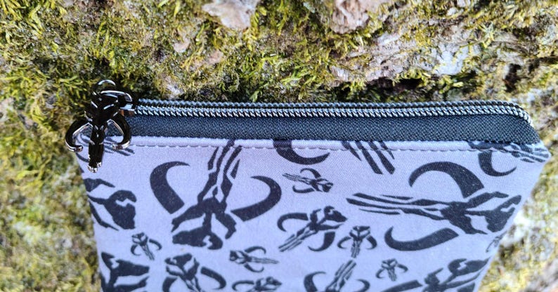 Puede incluir: Peque&ntilde;o estuche rectangular con cremallera negra y tirador plateado. El estuche es de color gris claro con un patr&oacute;n negro repetido de s&iacute;mbolos estilizados. El fondo es una superficie texturizada con musgo verde.