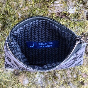 Puede incluir: Bolsa con cremallera abierta, con estampado gris y negro, con cremallera plateada y una peque&ntilde;a etiqueta azul en el interior que dice "GALACTIC SUMMER". La bolsa est&aacute; abierta, revelando un forro con estampado de hojas negras y grises.