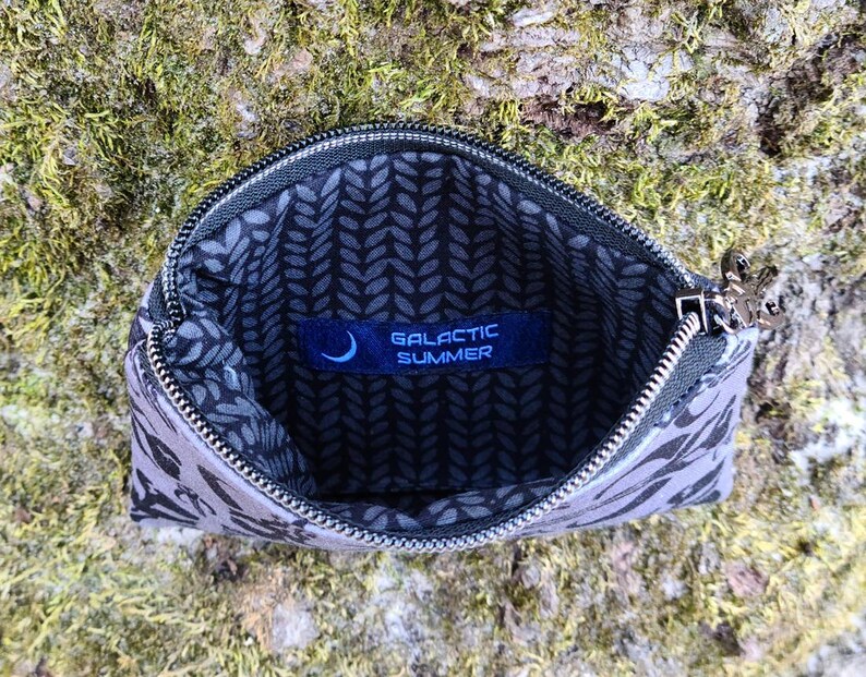 Puede incluir: Bolsa con cremallera abierta, con estampado gris y negro, con cremallera plateada y una peque&ntilde;a etiqueta azul en el interior que dice "GALACTIC SUMMER". La bolsa est&aacute; abierta, revelando un forro con estampado de hojas negras y grises.