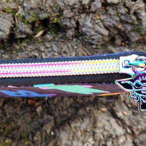 Puede incluir: Una bolsa negra con cremallera con un estampado abstracto colorido y un tirador de cremallera plateado con un personaje de dibujos animados.