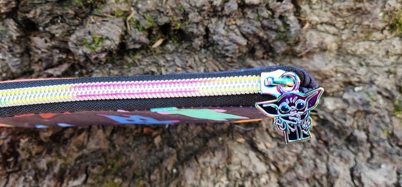 Puede incluir: Una bolsa negra con cremallera con un estampado abstracto colorido y un tirador de cremallera plateado con un personaje de dibujos animados.