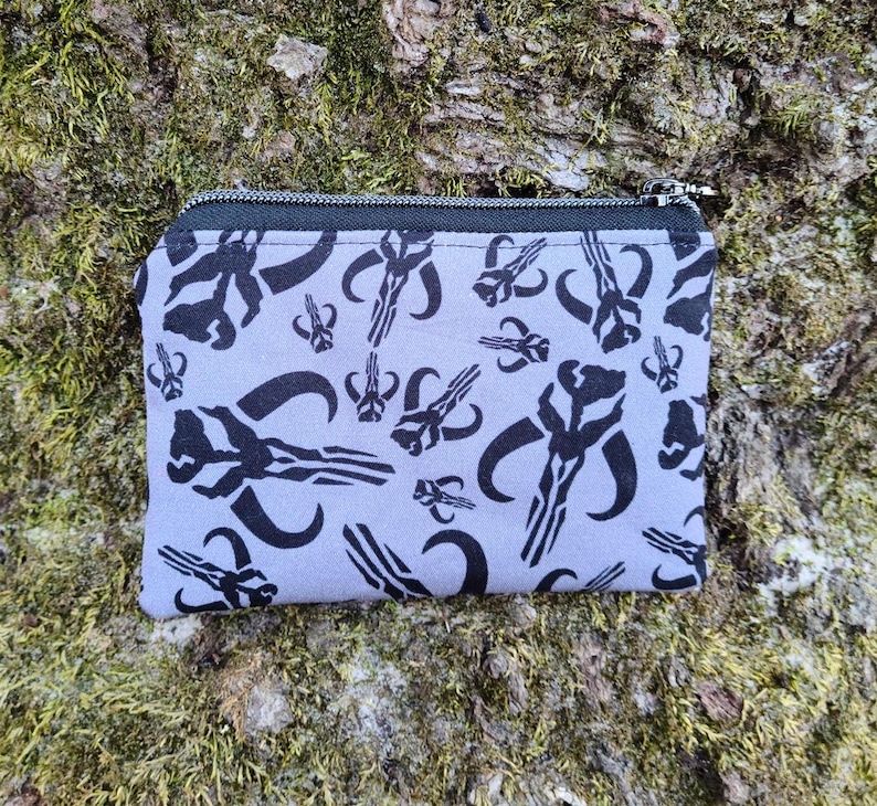 Puede incluir: Un peque&ntilde;o monedero rectangular con cierre de cremallera negro. La tela gris claro est&aacute; estampada con s&iacute;mbolos de calaveras mandalorianas negras. El monedero est&aacute; sobre un fondo de madera musgosa y texturizada.