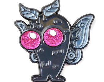 May include: A dark gray enamel pin featuring a stylized creature with large, glittery pink eyes and gray wings. The pin has a textured body and two leaf-shaped accents on top. The design is outlined in black.