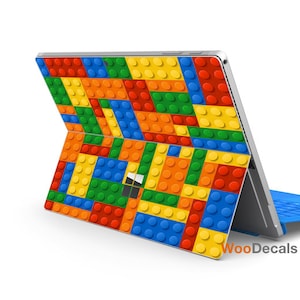 Może przedstawiać: Laptop z kolorowym wzorem w stylu Lego na pokrywie. Laptop jest zamknięty, a pokrywa skierowana jest w stronę aparatu. Laptop znajduje się na białym tle. W prawym dolnym rogu obrazu widoczny jest tekst "WooDecals".