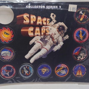 May include: A set of 10 Space Caps collector series 2 featuring astronaut patches with different designs. The patches are round and have a black background with colorful designs. The patches are displayed on a black background with the text "Space Caps" in white letters. The text "Collector Series 2" is also displayed in white letters.