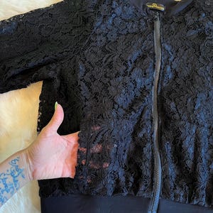 Può includere: Giacca bomber nera in pizzo con chiusura a zip e fascia nera sul fondo. La giacca ha un piccolo logo dorato che dice "Second Princess".