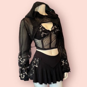 May include: Black sheer hooded top with lace detailing and a matching black skirt. The top has a cropped length and a fitted silhouette. The skirt is a-line and has a ruffled hem.