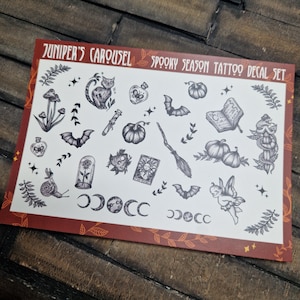 May include: A sheet of temporary tattoo decals with a spooky season theme. The sheet features various black and white illustrations, including a cat, bats, pumpkins, a witch's broom, and a fairy. The text "Juniper's Carousel Spooky Season Tattoo Decal Set" is printed at the top.