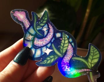 Pode incluir: Um adesivo holográfico com uma cobra rosa e azul enrolada em torno de uma videira verde com estrelas brancas. O adesivo tem um acabamento iridescente.