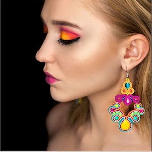 Pu&ograve; includere: Una donna con ombretto arancione e giallo brillante e rossetto rosa indossa grandi orecchini pendenti colorati. Gli orecchini sono realizzati con pi&ugrave; strati di filo e perline colorate, con una grande perla gialla in basso.