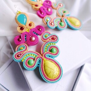 Pu&ograve; includere: Un paio di orecchini colorati con accenti turchese, rosa, giallo e verde. Gli orecchini sono realizzati con filo intrecciato e presentano grandi pietre a forma di goccia.