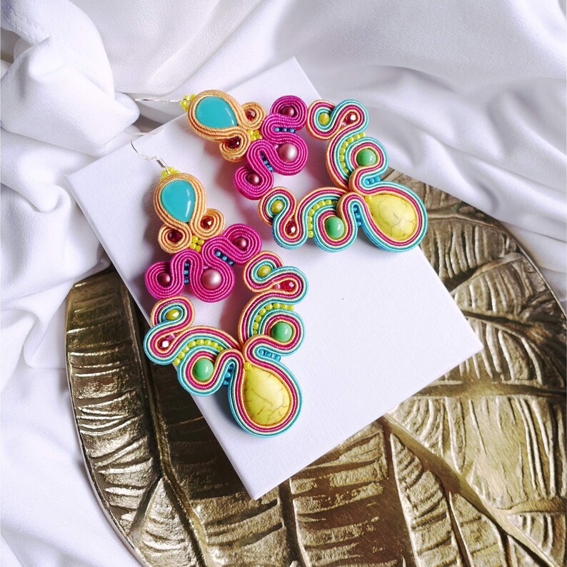 Pu&ograve; includere: Un paio di orecchini colorati con un design turchese, giallo, rosa e verde. Gli orecchini sono realizzati con un materiale intrecciato e hanno un motivo unico e intricato.