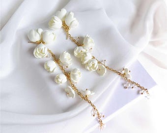 May include: A pair of elegant white floral earrings with gold chains and crystal accents. The earrings feature multiple white flower buds cascading down from the ear posts. The design is set against a white fabric background.
