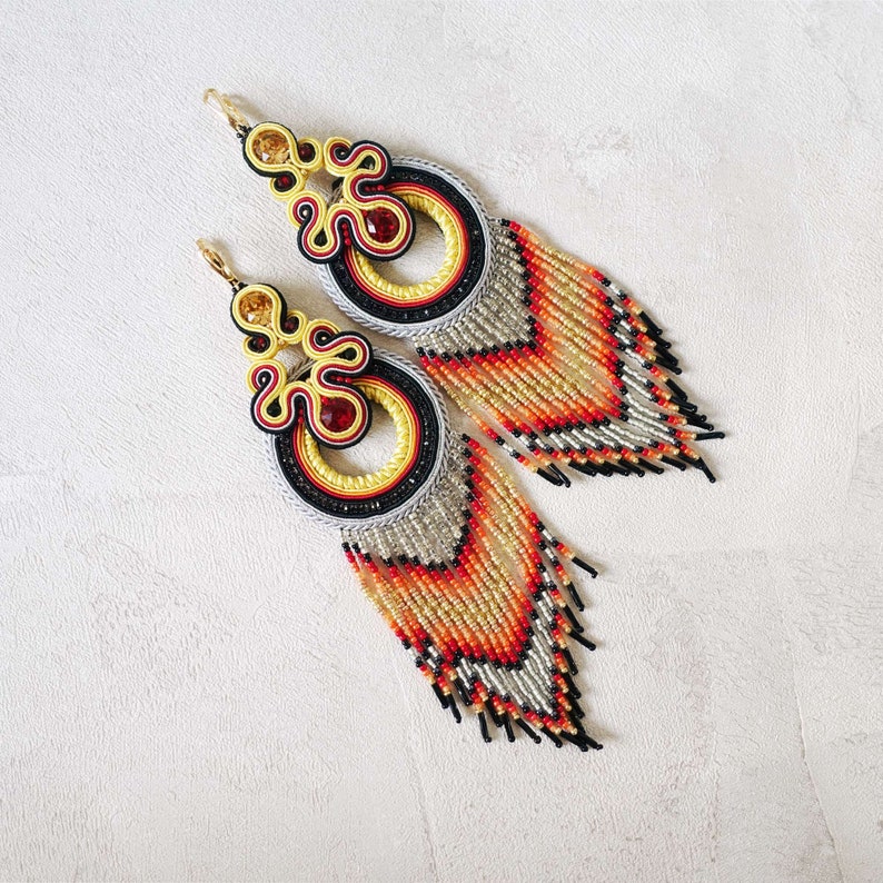 Op de afbeelding: Een paar lange, geperde oorbellen met een geweven ontwerp in geel, rood en zwart. De oorbellen hebben een franje van kralen in tinten oranje, geel en zwart.
