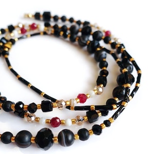 Op de afbeelding: Close-up van drie strengen kralenkettingen. De kettingen hebben zwarte, gouden, rode en heldere kralen. De kralen hebben verschillende vormen en maten, waaronder rond, vierkant en facetgeslepen. De kettingen zijn gelaagd en met elkaar verweven.