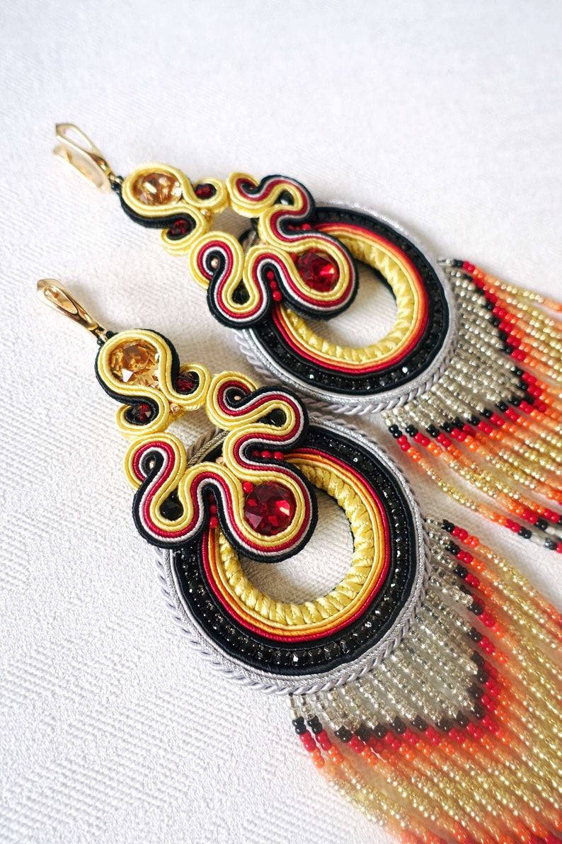 Op de afbeelding: Een paar lange, hangende oorbellen met een veelkleurig, ingewikkeld ontwerp. De oorbellen hebben een groot, rond, geel, rood en zwart pareldeel met een kleiner, driehoekig deel erboven. De oorbellen zijn afgewerkt met een lange, parelversierde franje in tinten oranje, rood, zwart en goud.