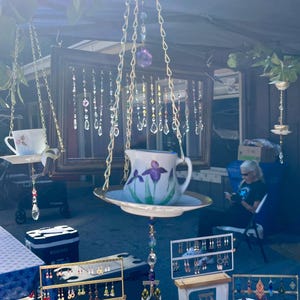 May include: An outdoor market scene featuring hanging teacups and saucers adorned with floral designs. The teacups are suspended by chains and embellished with crystals. Various earring displays are visible, showcasing a variety of styles and colors.