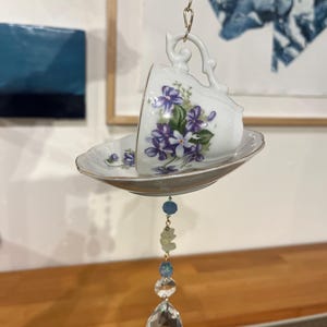 以下が含まれることがあります： 紫色の花と緑の葉のフローラルデザインが施された、白いティーカップとソーサーのセット。 ティーカップは金のチェーンで吊り下げられており、下にはクリスタルとビーズの装飾ペンダントが付いています。 ティーカップは傾いています。