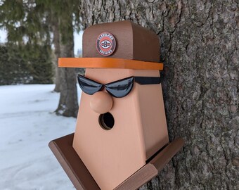 Może przedstawiać: Drewniany domek dla ptaków w kształcie twarzy noszącej okulary przeciwsłoneczne i brązowy kapelusz z logo Cleveland Browns. Domek dla ptaków ma mały otwór, przez który ptaki mogą wejść.