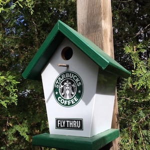 Könnte beinhalten: Ein grünes und weißes Vogelhaus mit einem Starbucks-Logo und den Worten "Fly Thru" auf der Vorderseite. Das Vogelhaus ist an einem Holzpfosten befestigt.