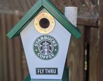 Puede incluir: Una casita de pájaros verde y blanca con un logotipo de Starbucks y las palabras "Fly Thru" en la parte delantera. La casita tiene una pequeña chimenea y una abertura redonda para que los pájaros puedan entrar.