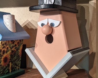 Puede incluir: Una casita de pájaros de madera con forma de persona que lleva una gorra de béisbol negra con un logotipo "NY" blanco. La casita de pájaros tiene una cara rosa con una nariz grande y redonda, ojos azules y una boca abierta de par en par. La casita de pájaros está sentada en un estante de madera.