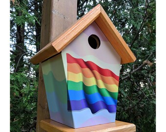 May include: A wooden birdhouse painted with a rainbow flag design. The birdhouse has a small opening for birds to enter and a wooden perch.