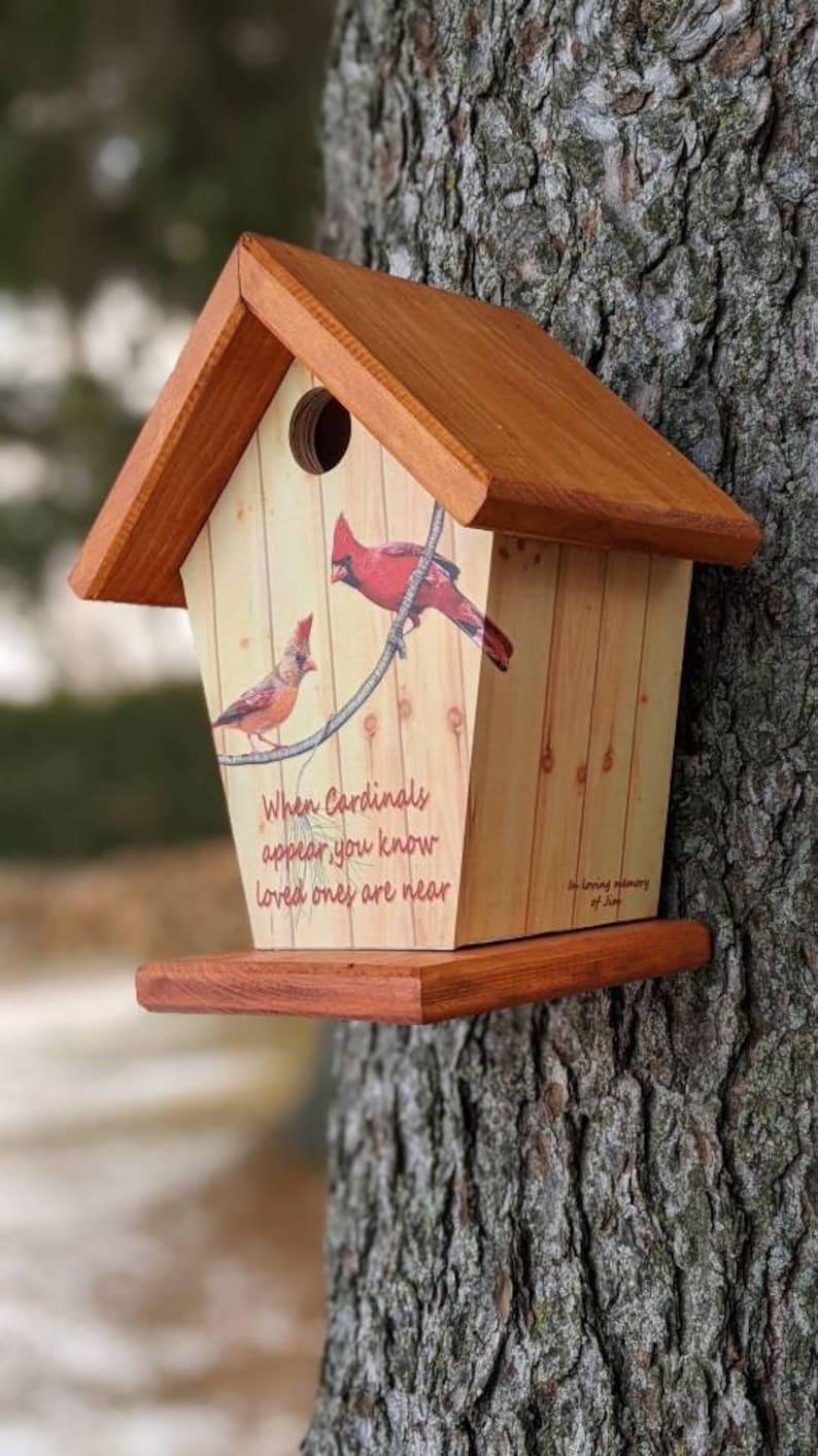 May include: Wooden birdhouse with a pitched roof, featuring a painted design of two cardinals perched on a wire. The birdhouse has a small round entrance hole and a wooden perch. Text on the front reads "When Cardinals appear, you know loved ones are near."