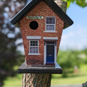 May include: A small birdhouse shaped like a brick house with a black roof and a blue door. The birdhouse has a sign that says "Stanbridge St."