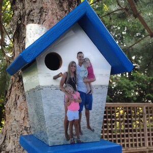 May include: A blue birdhouse with a photo of a family on a beach. The photo shows a man, a woman, and a young girl. The birdhouse has a blue roof and a white base.