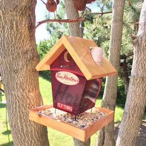 May include: A wooden bird feeder shaped like a Tim Hortons coffee cup with a red and white logo and the words "Fly Thru". The feeder is attached to a tree with two rusty metal birds perched on a branch above it.