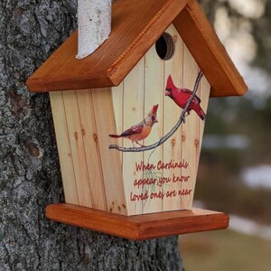 May include: Wooden birdhouse with a pitched roof and a small chimney. The birdhouse is decorated with images of red cardinals perched on a branch, with the text "When Cardinals appear you know loved ones are near."