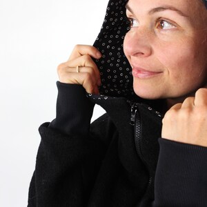 May include: A black hooded sweatshirt with a white polka dot lining. The hood is pulled up and the person is looking to the right.