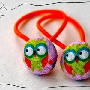 Könnte beinhalten: Zwei orangefarbene Haargummis mit Knöpfen aus lila und grünem Eulenstoff. Die Eulen haben große Augen und einen leuchtend orangefarbenen Schnabel.
